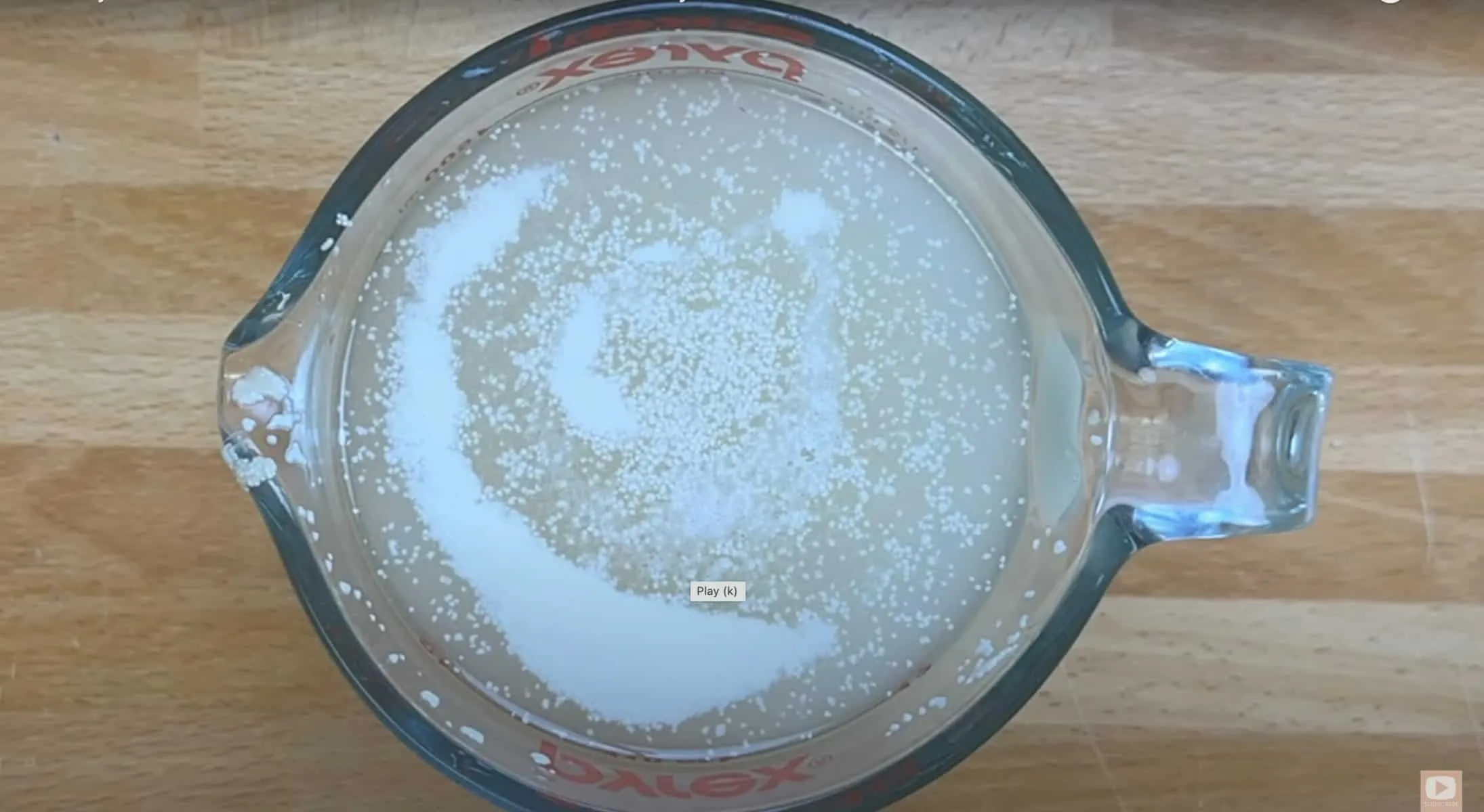 Yeast blooming in warm water inside a Pyrex measuring cup on a wooden surface.